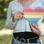 An adult holding a rainbow heart, symbolizing LGBTQ pride, outdoors.