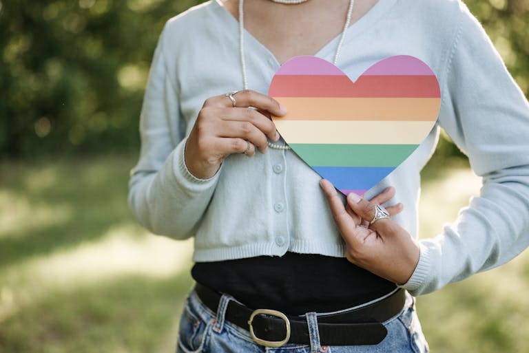 An adult holding a rainbow heart, symbolizing LGBTQ pride, outdoors.